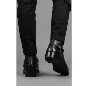 May include: Black leather ankle boots with a low heel and a strap with a silver buckle. The boots are paired with black cargo pants. The image is shot from a low angle, highlighting the boots' design and the texture of the leather.