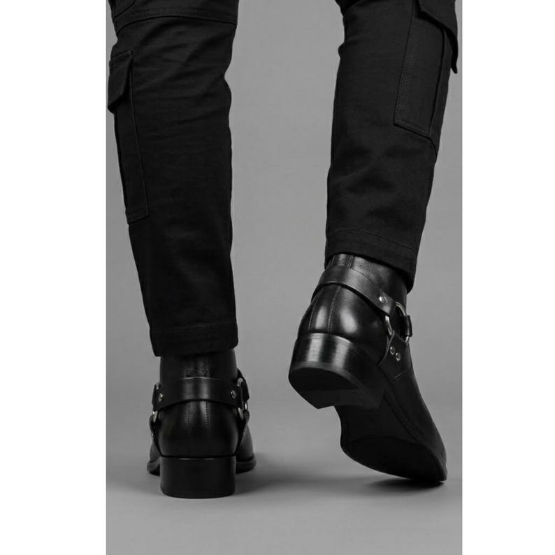 May include: Black leather ankle boots with a low heel and a strap with a silver buckle. The boots are paired with black cargo pants. The image is shot from a low angle, highlighting the boots' design and the texture of the leather.