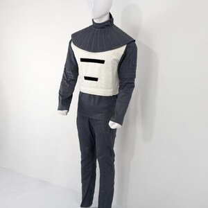 Może przedstawiać: Futurystyczny kostium wystawiony na manekinie. Str&oacute;j obejmuje ciemnoszarą kurtkę z wysokim kołnierzem, kremową kamizelkę z dwoma czarnymi poziomymi paskami oraz pasujące ciemnoszare spodnie. Manekin ma białą głowę i dłonie.