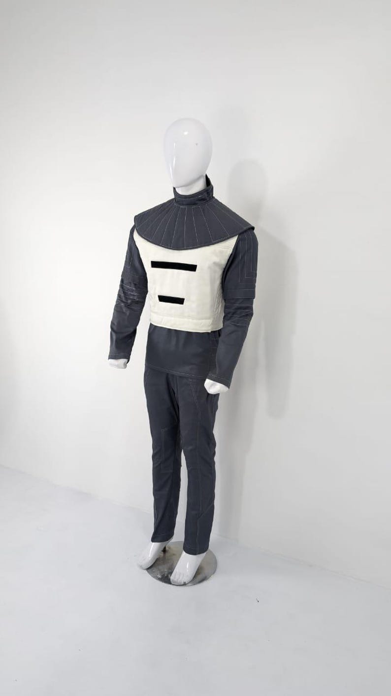 Może przedstawiać: Futurystyczny kostium wystawiony na manekinie. Str&oacute;j obejmuje ciemnoszarą kurtkę z wysokim kołnierzem, kremową kamizelkę z dwoma czarnymi poziomymi paskami oraz pasujące ciemnoszare spodnie. Manekin ma białą głowę i dłonie.