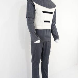 Może przedstawiać: Futurystyczny str&oacute;j na manekinie. Zestaw zawiera ciemnoszarą koszulę z długim rękawem, pasujące spodnie i białą kamizelkę z czarnymi poziomymi paskami. Str&oacute;j ma wysoki, pikowany kołnierz i białe szwy.