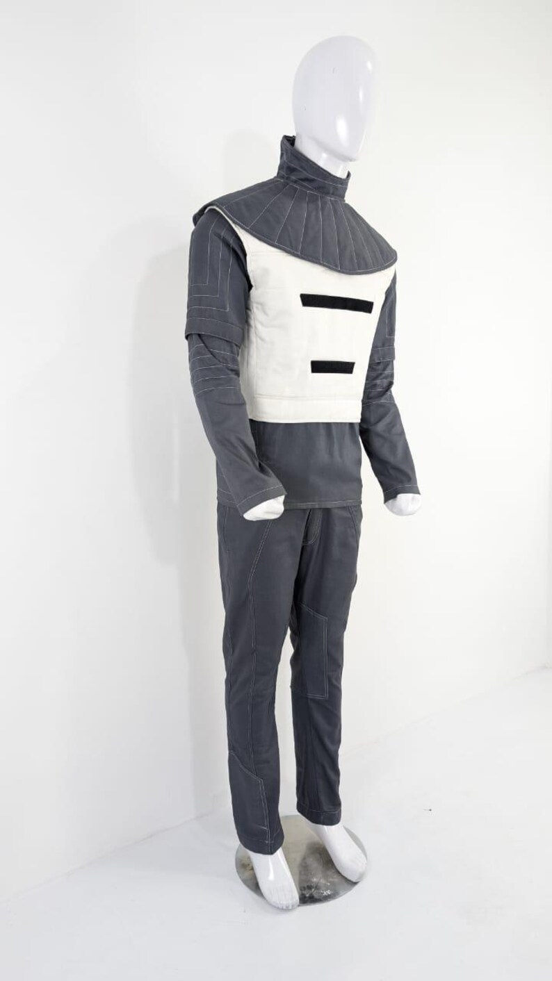 Może przedstawiać: Futurystyczny str&oacute;j na manekinie. Zestaw zawiera ciemnoszarą koszulę z długim rękawem, pasujące spodnie i białą kamizelkę z czarnymi poziomymi paskami. Str&oacute;j ma wysoki, pikowany kołnierz i białe szwy.