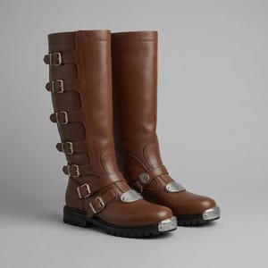 May include: A pair of brown leather knee-high boots. Each boot features multiple silver buckles and a silver plate on the toe and ankle strap. The boots have a black sole and a stacked heel.