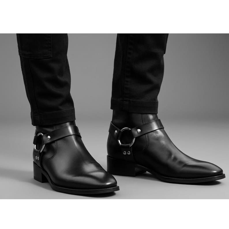 May include: Black leather ankle boots with a harness strap and silver-tone hardware. The boots have a low heel and a pointed toe. The boots are worn with black pants.