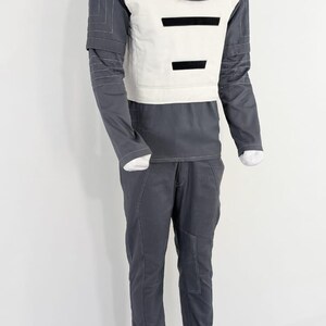 Może przedstawiać: Futurystyczny kostium wystawiony na manekinie. Str&oacute;j składa się z szarej g&oacute;ry z wysokim kołnierzem, białej kamizelki i czarnych akcent&oacute;w. Pasujące szare spodnie z białymi przeszyciami dopełniają całość. Kostium ma elegancką, nowoczesną estetykę.