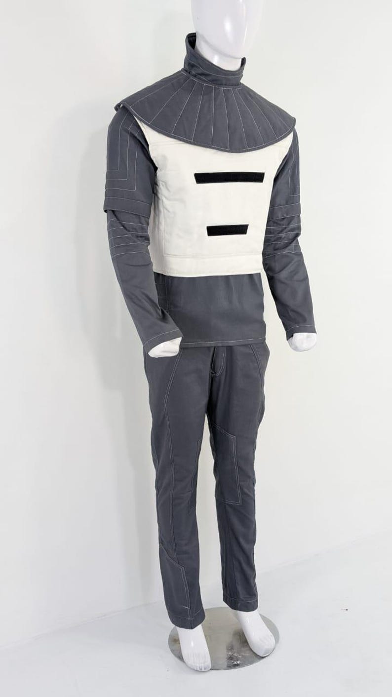 Może przedstawiać: Futurystyczny kostium wystawiony na manekinie. Str&oacute;j składa się z szarej g&oacute;ry z wysokim kołnierzem, białej kamizelki i czarnych akcent&oacute;w. Pasujące szare spodnie z białymi przeszyciami dopełniają całość. Kostium ma elegancką, nowoczesną estetykę.