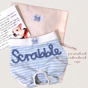 Puede incluir: Ropa interior para perros a rayas azules y blancas con la palabra "Scrabble" bordada en azul. La ropa interior tiene una cintura blanca y una etiqueta "Butt Hugger". También hay una bolsa beige con cordón y una tarjeta con una ilustración de perro.