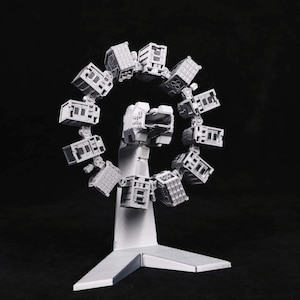 May include: A white, 3D-printed model of a Ferris wheel-like structure. The wheel is composed of small, rectangular cabins. The base is a white, angular support structure, set against a black background.