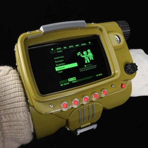 May include: A green and black Pip-Boy wrist-mounted device with a screen displaying game stats. The device has red buttons and a handle. The screen shows the text "STAT INV DATA MAP RADIO SPECIAL PERKS Animal Friend Big Leagues Bloody Mess Pickpocket Sniper".