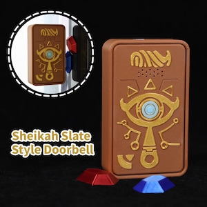 Puede incluir: Un timbre de puerta marrón estilo Sheikah Slate con detalles dorados y un diseño de ojo azul. El timbre rectangular incluye el texto "Sheikah Slate Style Doorbell". Dos gemas decorativas en la base.