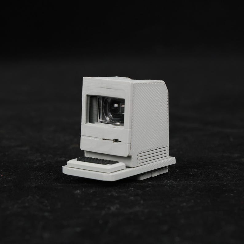 May include: A miniature, off-white computer with a small screen and a black keyboard. The computer has a classic design, with a rectangular body and a built-in screen. The keyboard is a small, rectangular shape.