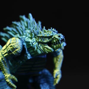 Può includere: Un primo piano di una statuetta di mostro verde e blu. La figura ha un aspetto strutturato e squamoso con pinne dorsali affilate e una bocca minacciosa. L'illuminazione evidenzia gli intricati dettagli della scultura su uno sfondo scuro.