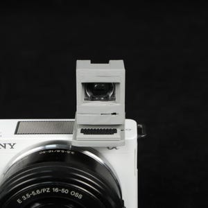 May include: A gray, miniature computer-shaped viewfinder attached to a white camera. The viewfinder has a small screen and a keyboard design. The camera lens is black, and the camera body is white. The background is black.