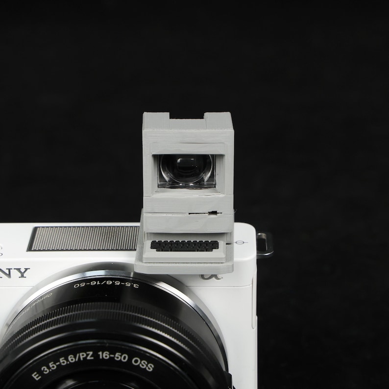 May include: A gray, miniature computer-shaped viewfinder attached to a white camera. The viewfinder has a small screen and a keyboard design. The camera lens is black, and the camera body is white. The background is black.