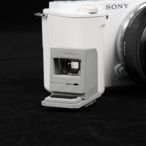May include: A gray, miniature computer-shaped viewfinder attached to a white Sony camera. The viewfinder has a small keyboard and screen, resembling a vintage computer. The camera lens is black.