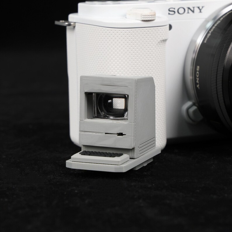May include: A gray, miniature computer-shaped viewfinder attached to a white Sony camera. The viewfinder has a small keyboard and screen, resembling a vintage computer. The camera lens is black.