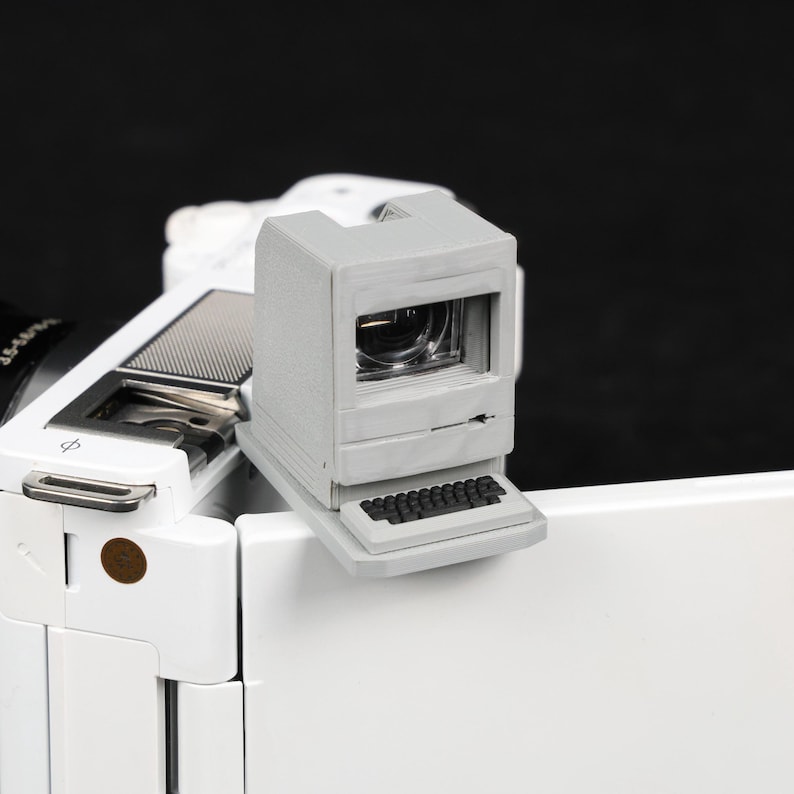 May include: A gray, miniature computer-shaped viewfinder attached to a white camera. The viewfinder has a small keyboard at the bottom and a screen-like opening. The camera body is white with black accents, set against a black background.