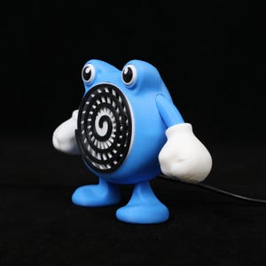 Pode incluir: Uma figura azul com luvas e p&eacute;s brancos, e olhos grandes em preto e branco. A figura tem um desenho em espiral no peito e um cabo preto que se estende da sua base. O design geral &eacute; de desenho animado.