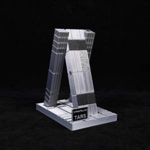 May include: A gray and black model of the TARS robot from the movie Interstellar. The model is made of rectangular blocks and has the word "TARS" printed on it. The model is on a rectangular base with the words "Interstellar TARS" printed on it.