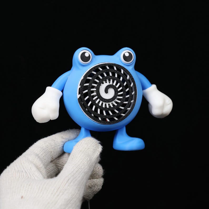 Pode incluir: Uma figura azul, semelhante a um desenho animado, com um design em espiral no centro. A figura tem luvas e p&eacute;s brancos e &eacute; segurada por uma m&atilde;o enluvada contra um fundo preto. A figura tem olhos grandes e redondos.