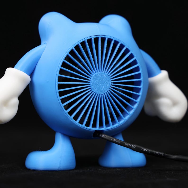 Pode incluir: Um ventilador azul de novidade em forma de personagem de desenho animado com um ventilador redondo no centro. O ventilador tem luvas e p&eacute;s brancos. Um cabo de alimenta&ccedil;&atilde;o preto se estende da parte inferior do ventilador.