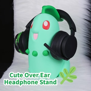 Op de afbeelding: Een mintgroene hoofdtelefoonstandaard in de vorm van een stripfiguur, met rode ogen en een lachende roze mond. Zwarte over-ear hoofdtelefoons rusten op de standaard. De tekst "Cute Over Ear Headphone Stand" staat onderaan.