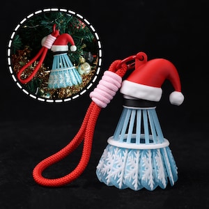May include: A novelty Christmas decoration in the shape of a badminton shuttlecock, adorned with a red Santa hat and snowflake patterns. The shuttlecock is light blue with white snowflake details, and has a red lanyard. A pink cord wraps the lanyard.