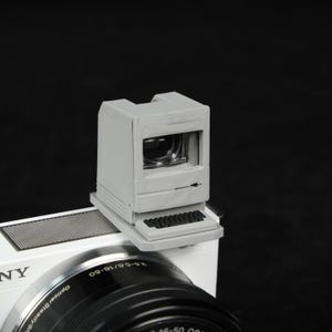 May include: A gray, 3D-printed camera viewfinder shaped like a vintage computer. The viewfinder is mounted on a white camera body with a black lens. The design includes a miniature keyboard and screen, evoking a retro aesthetic.