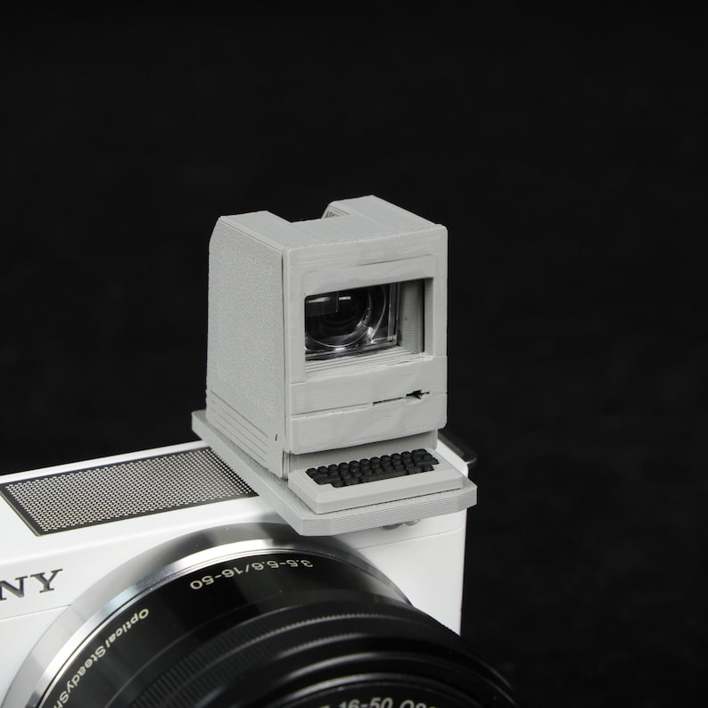 May include: A gray, 3D-printed camera viewfinder shaped like a vintage computer. The viewfinder is mounted on a white camera body with a black lens. The design includes a miniature keyboard and screen, evoking a retro aesthetic.