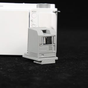 May include: A gray 3D-printed camera viewfinder attachment. The attachment is rectangular with a small, dark opening and a textured surface. It is mounted on a white camera body with buttons and a menu dial.