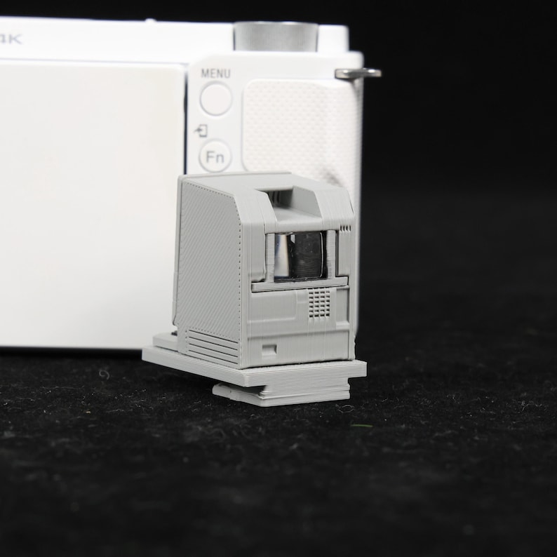 May include: A gray 3D-printed camera viewfinder attachment. The attachment is rectangular with a small, dark opening and a textured surface. It is mounted on a white camera body with buttons and a menu dial.