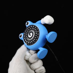 Pode incluir: Uma figura azul, semelhante a um desenho animado, com um design em espiral na frente. Tem dois olhos, dois p&eacute;s e uma luva branca. A figura &eacute; segurada contra um fundo preto.