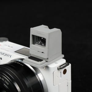 May include: A gray, 3D-printed camera viewfinder accessory shaped like a vintage computer, mounted on a white Sony camera. The accessory has a small keyboard detail and a screen-like opening. The camera lens is visible below.