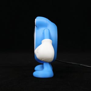 Pode incluir: Uma figura azul semelhante a um desenho animado com luvas e p&eacute;s brancos. A figura tem um corpo arredondado e um pequeno rosto circular. Um cabo preto estende-se da parte inferior da figura.