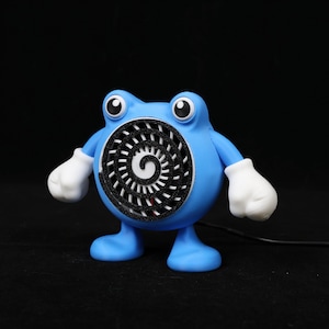 Pode incluir: Uma figura azul, semelhante a um desenho animado, com um design em espiral na frente. A figura tem luvas brancas e olhos grandes. O fundo &eacute; preto.