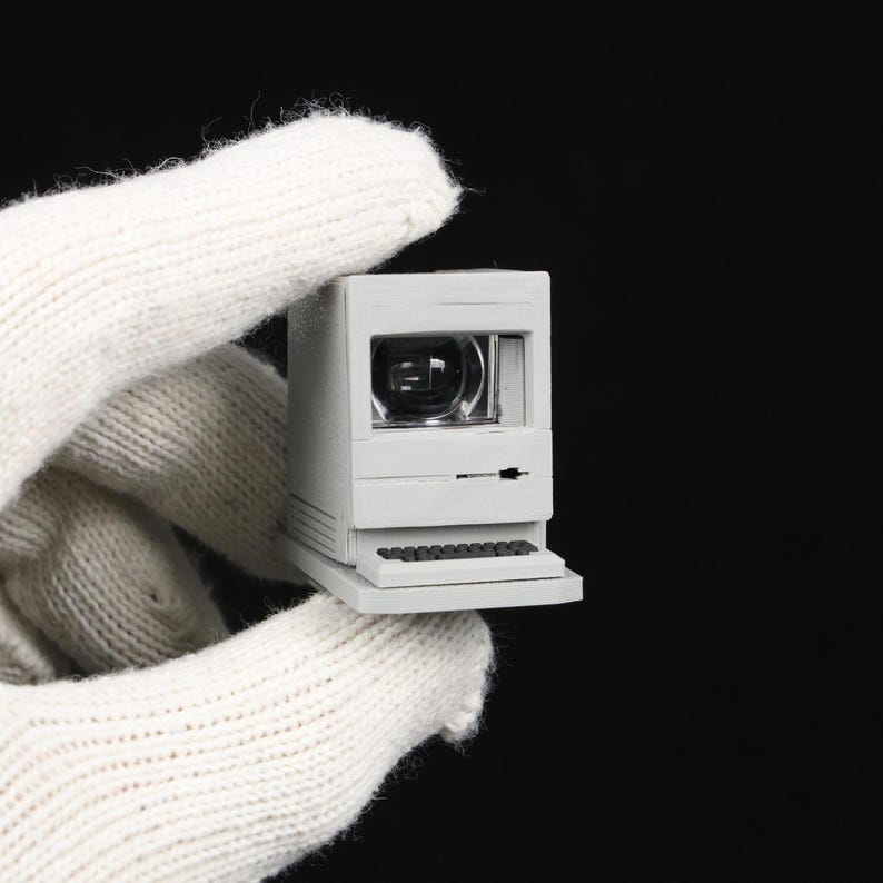 May include: A miniature, off-white computer with a screen and keyboard. The small computer has a retro design, reminiscent of older desktop computers. The image is set against a black background.