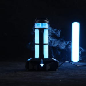 Puede incluir: Un dispositivo electrónico negro y azul con un diseño futurista. El dispositivo tiene una base rectangular con un patrón geométrico y una torre central con dos luces azules verticales. Una flecha blanca apunta hacia abajo. A la derecha hay un tubo de luz azul.