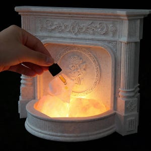 Może przedstawiać: Biały, dekoracyjny kominek z motywem róży pośrodku. Kominek wypełniony jest podświetlanymi różowymi kamieniami solnymi. Ręka trzyma zakraplacz z żółtym płynem, dodając go do kamieni.