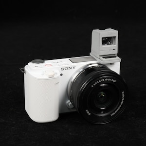 May include: A white Sony digital camera with a black lens and a miniature gray computer attached to the top. The camera has a textured grip and a red button. The lens has the specifications: E 3.5-5.6/PZ 16-50 OSS.
