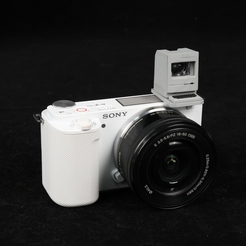 May include: A white Sony digital camera with a black lens and a miniature gray computer attached to the top. The camera has a textured grip and a red button. The lens has the specifications: E 3.5-5.6/PZ 16-50 OSS.