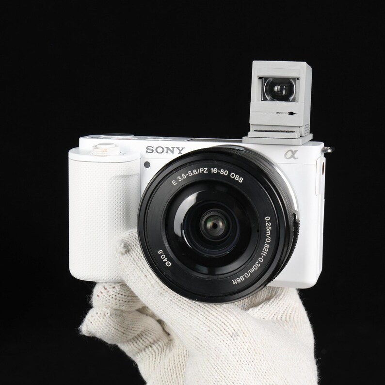 May include: A white Sony digital camera with a black lens and a small, gray accessory on top. The lens has the text "E 3.5-5.6/PZ 16-50 OSS" and "&alpha;" printed on it. The camera is held by a gloved hand against a black background.
