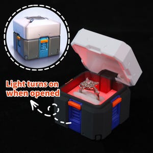 May include: A black, blue, and orange box-shaped ring holder with a white lid. The box is open, revealing a silver ring with a decorative design. The interior is lit with red light. Text on the image reads "Light turns on when opened."