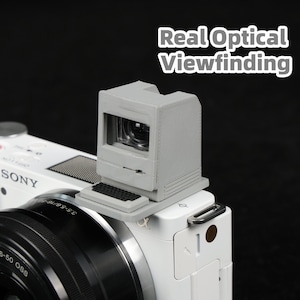 Peut inclure: Un viseur gris en forme d'écran d'ordinateur rétro est monté sur un appareil photo Sony blanc. Le texte "Real Optical Viewfinding" est affiché au-dessus du viseur. L'objectif de l'appareil photo est noir.