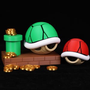 May include: A green and red turtle shell toy with a white trim, a green pipe, and gold coins on a brown brick platform. The shells are from a video game.