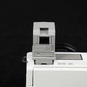 May include: A gray camera flash attachment mounted on a white camera body. The flash has a rectangular design with a central black screen and a grid-like pattern. The camera body has the text "ZV-E10 4K".