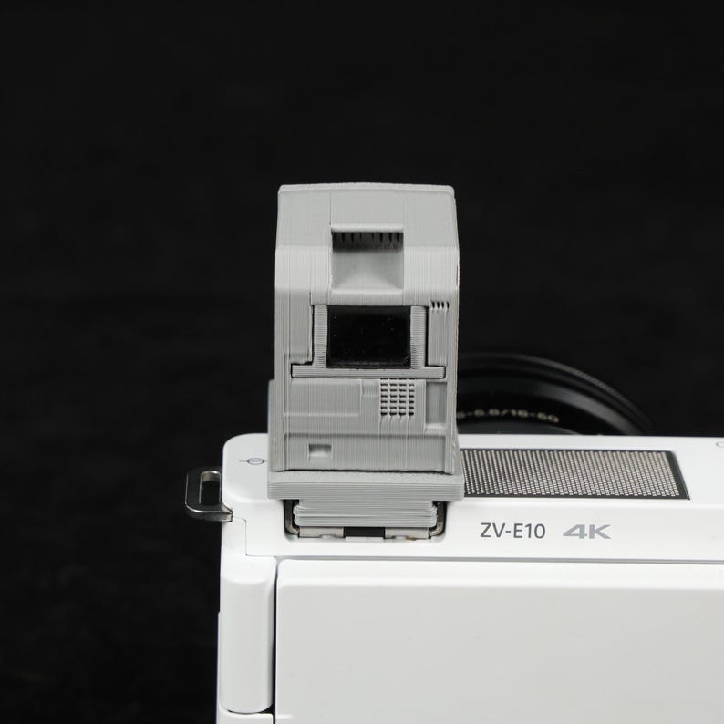 May include: A gray camera flash attachment mounted on a white camera body. The flash has a rectangular design with a central black screen and a grid-like pattern. The camera body has the text "ZV-E10 4K".
