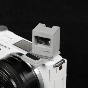 May include: A gray viewfinder attached to a white Sony camera. The viewfinder has a rectangular shape with a small screen and a keyboard-like detail. The camera lens is black with silver accents.