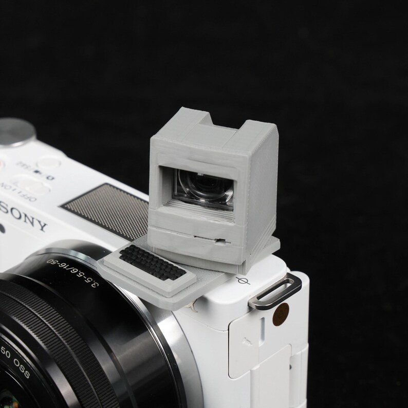 May include: A gray viewfinder attached to a white Sony camera. The viewfinder has a rectangular shape with a small screen and a keyboard-like detail. The camera lens is black with silver accents.