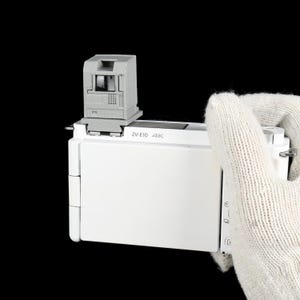 May include: A white digital camera with a gray viewfinder attachment. The camera has the text "ZV-E10 4K" printed on it. The camera is held by a gloved hand against a black background.