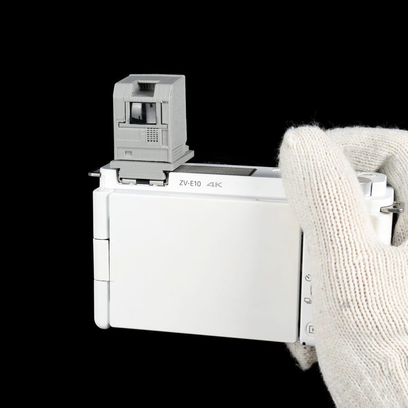 May include: A white digital camera with a gray viewfinder attachment. The camera has the text "ZV-E10 4K" printed on it. The camera is held by a gloved hand against a black background.